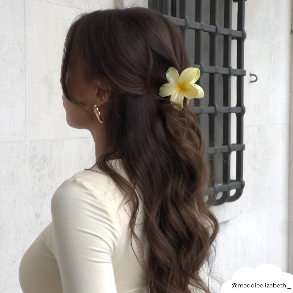 Woman with long wavy brown hair wearing a butter yellow tropical flower claw clip, facing a light wall.