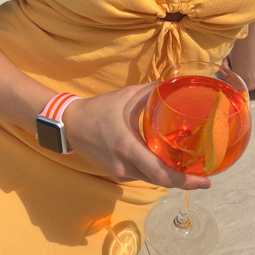 Person holding a glass of orange cocktail with a lemon slice, wearing a yellow dress and apple watch on wrist with pink and orange watch strap.