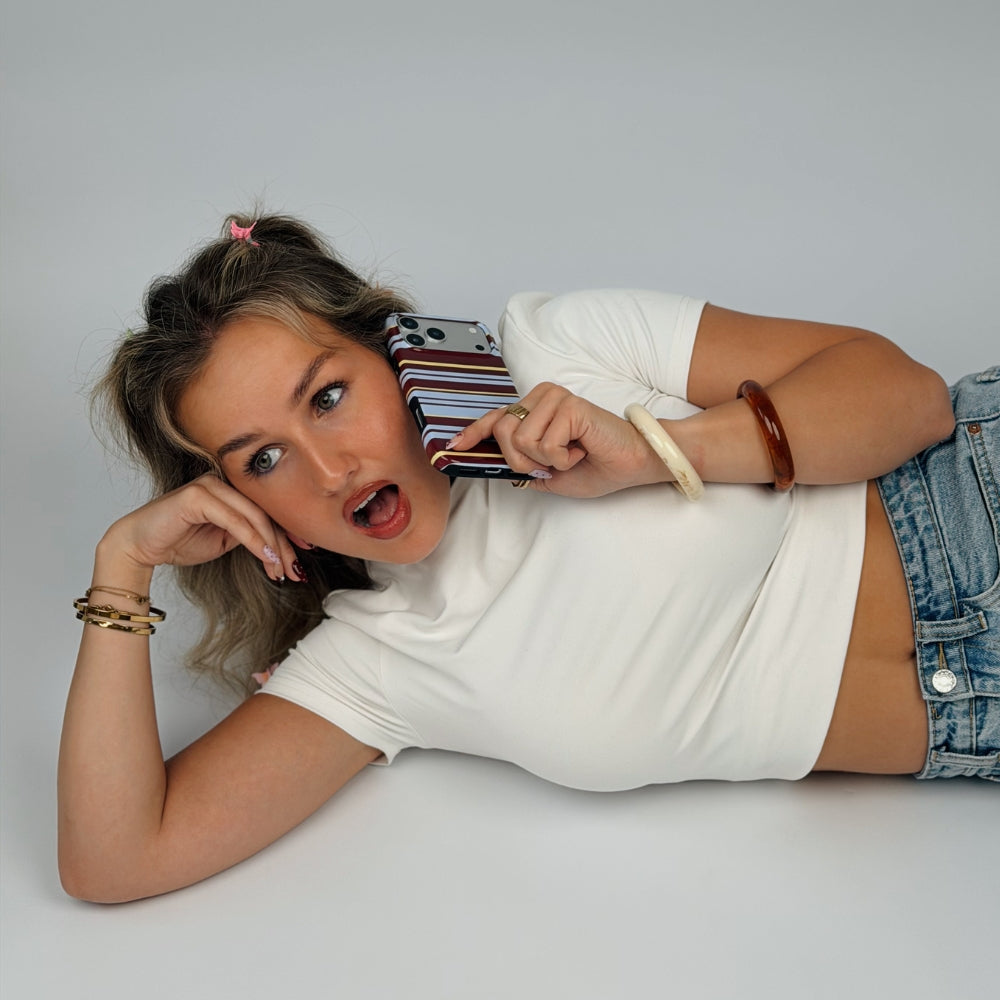 A woman lying on a light grey floor wearing a white short-sleeve crop top and blue denim shorts, holding the Mary-Kate striped phone case to her ear with one hand while resting her head on her elbow with the other.