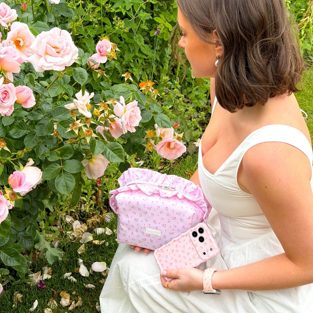 Woman holding ditsy floral pink ruffles makeup bag and matching phone case and watch strap, in front of a floral background