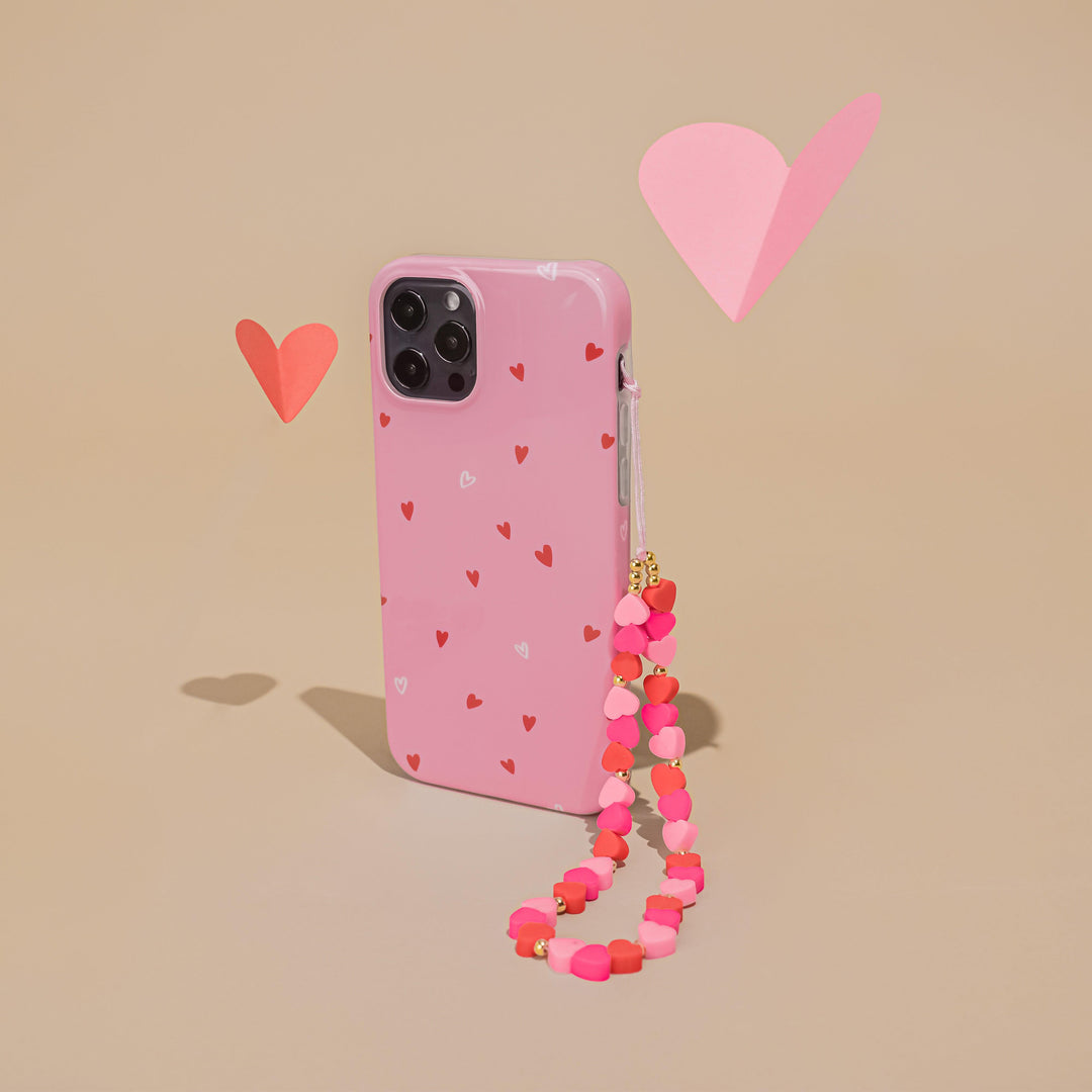 A pink phone case with small red and white hearts, a pink and red beaded strap, and pink heart-shaped paper cutouts floating around, on a beige background.