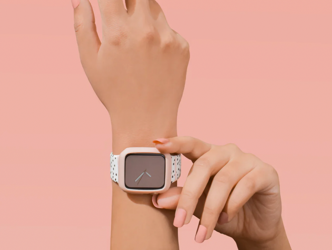 A hand with manicured nails wears a smartwatch with a pink case, a polka-dot strap, and a minimalist dark face displaying two clock hands. The background is light pink.