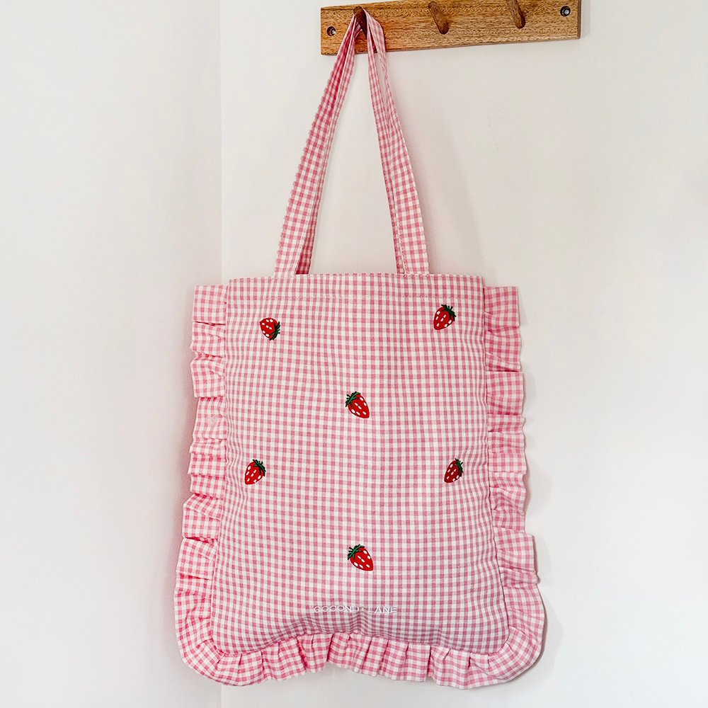 A pink gingham tote bag with ruffled edges and small strawberry designs hangs on a wooden hook against a white wall.