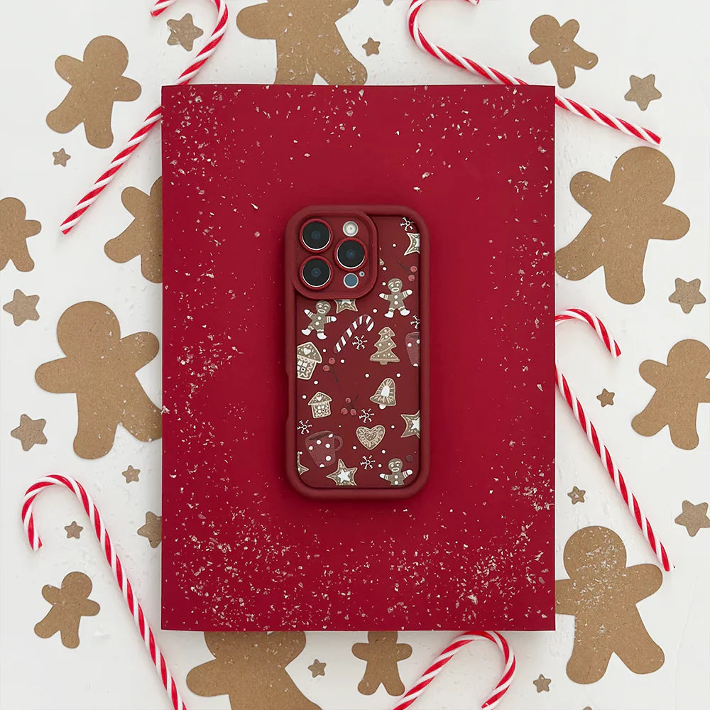 A phone with a red case decorated with festive gingerbread and holiday icons is placed on a red surface, surrounded by gingerbread cut-outs and candy canes. White sprinkles add a snowy effect.