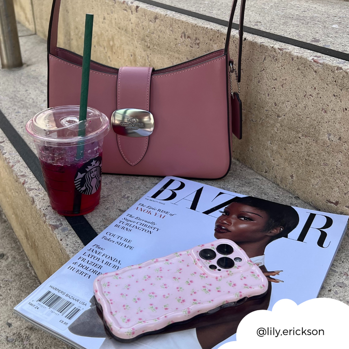 A pink handbag, a Starbucks drink with a green straw, a Harper's Bazaar magazine featuring a woman on the cover, and Coconut Lane's Wavy Phone Case in Ditsy Floral Pink are placed on concrete steps. The Instagram handle @lily.erickson is positioned in the bottom right corner within a white cloud shape.