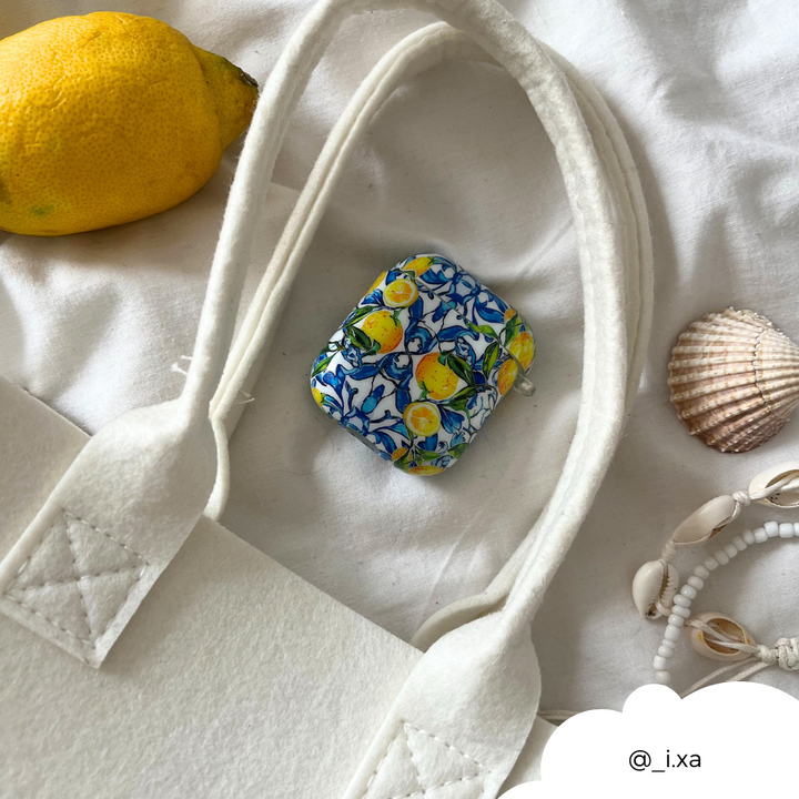 The vibrant AirPods Case - Amalfi Lemon from Coconut Lane, featuring premium materials and IMD Print Technology, is displayed on a white surface next to a white bag strap, a lemon, seashells, and a white beaded bracelet. In the lower right corner, there is a cloud-shaped cutout with the text "@_i_xa".