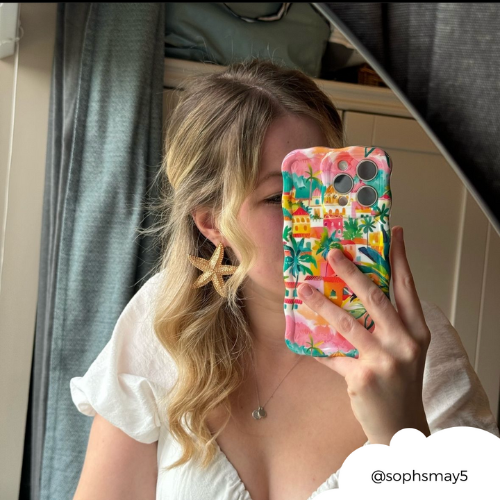 A person with wavy blond hair wearing a white top and starfish-shaped earrings is taking a mirror selfie with their smartphone. The phone case, an eye-catching and colorful design from Coconut Lane called the Wavy Phone Case - Portofino Dreams, features a beautiful Italian seaside motif. The Instagram handle "@sophsmay5" is visible in the bottom right corner.