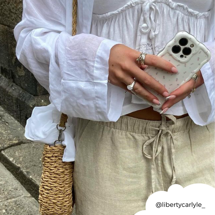 A person wearing a white long-sleeve blouse and beige drawstring pants holds an iPhone adorned with the Wavy Phone Case - Ditsy Floral White from Coconut Lane in both hands. They have painted nails and wear rings. A woven straw bag is slung over their shoulder, and their Instagram handle, @libertycarlyle_, is at the bottom right corner.