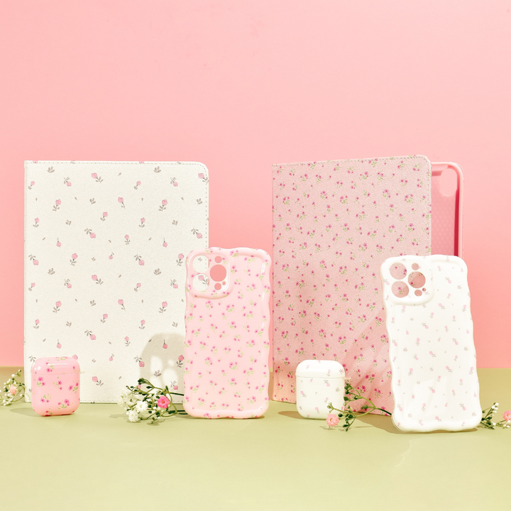 A display of floral-themed tech accessories includes two tablet cases, four iPhone cases (featuring the Wavy Phone Case - Ditsy Floral White by Coconut Lane), and two AirPods cases. The items are arranged on a light green surface against a pink backdrop, with small white flowers accentuating the display.