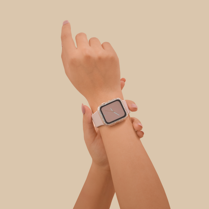 A close-up of a person's hands against a beige background. The person is wearing an Apple Watch with a white band on their left wrist. Their right hand, adorned with the Coconut Lane 2-in-1 Apple Watch Case & Built-In Screen Protector - Clear Diamante, is adjusting the watch, pointing upwards. The smartwatch display shows an analog clock face.