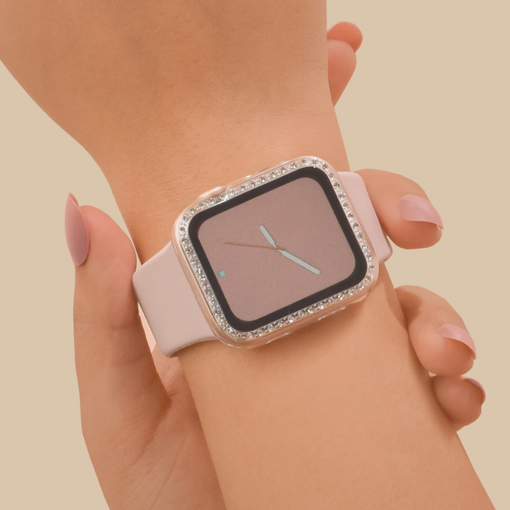 A person wearing an Apple Watch with a pink strap and a square, brown face, adorned with Coconut Lane's 2-in-1 Apple Watch Case & Built-In Screen Protector in Clear Diamante. The glittery border around the screen resembles the elegance of a diamante watch case. The person's nails are neatly manicured as they touch the device with their other hand against a beige background.