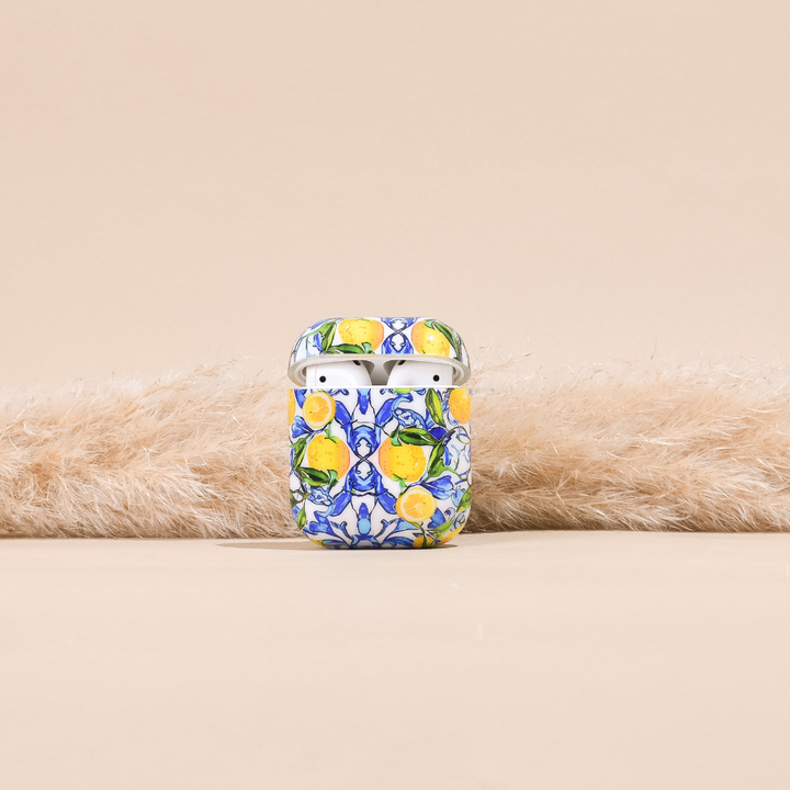 The AirPods Case - Amalfi Lemon by Coconut Lane, crafted from premium materials, showcases a vibrant floral and lemon pattern. It rests on a soft, white, fluffy surface against a neutral beige background. The top of the case is slightly open, revealing the white AirPods inside.