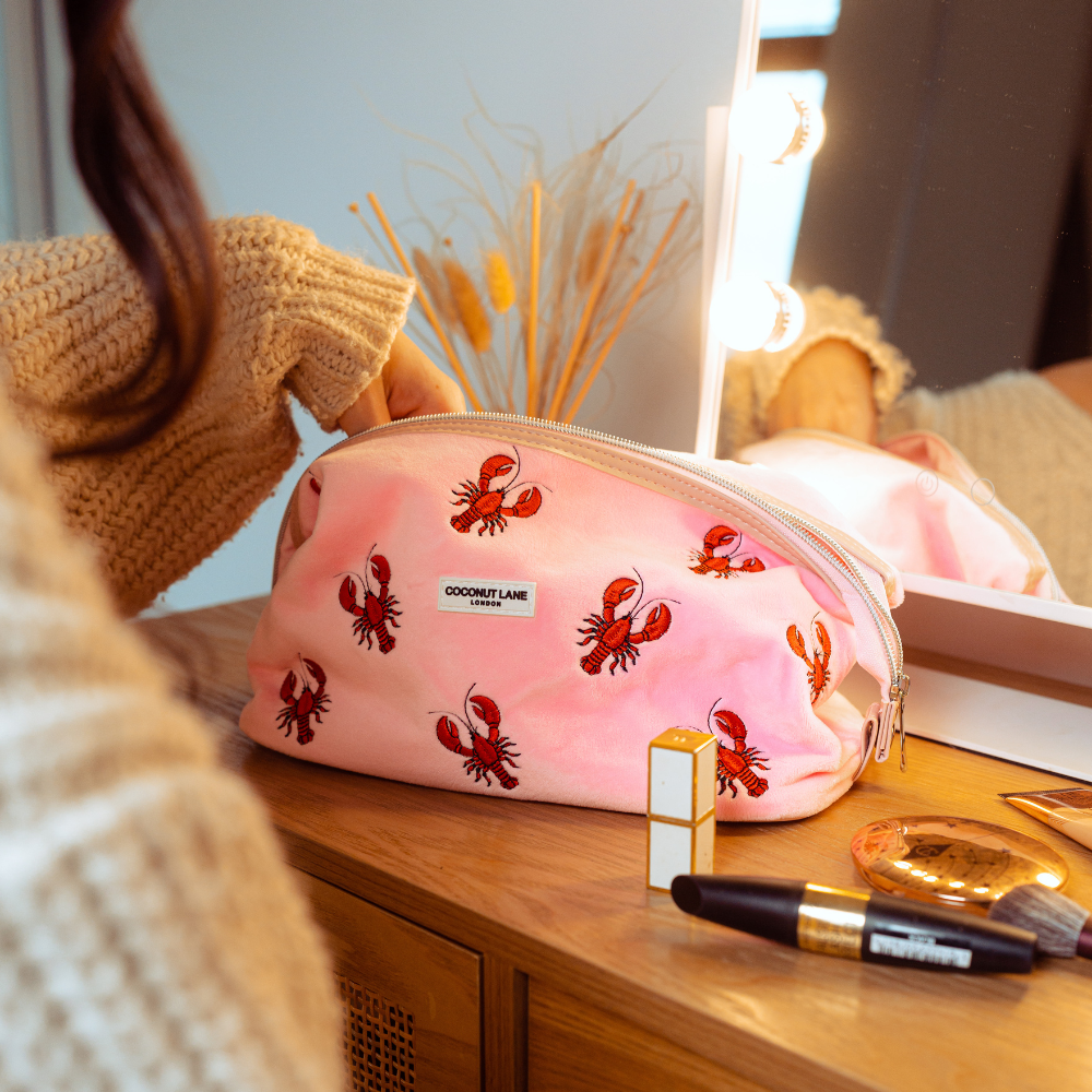 Hand reaching into pink velvet expandable makeup bag with lobster design, on vanity near cosmetics and a lit mirror.