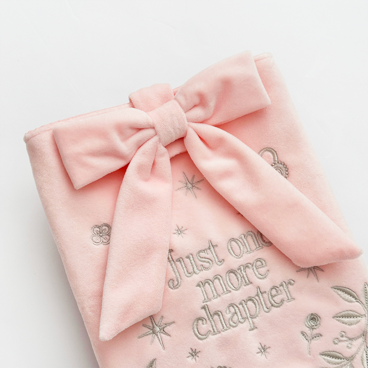 Book Sleeve made from soft blush pink velvet with grey embroidered floral details, stars and 'Just One More Chapter' lettering, finished with a large pink velvet bow and Coconut Lane logo, photographed on a white background.