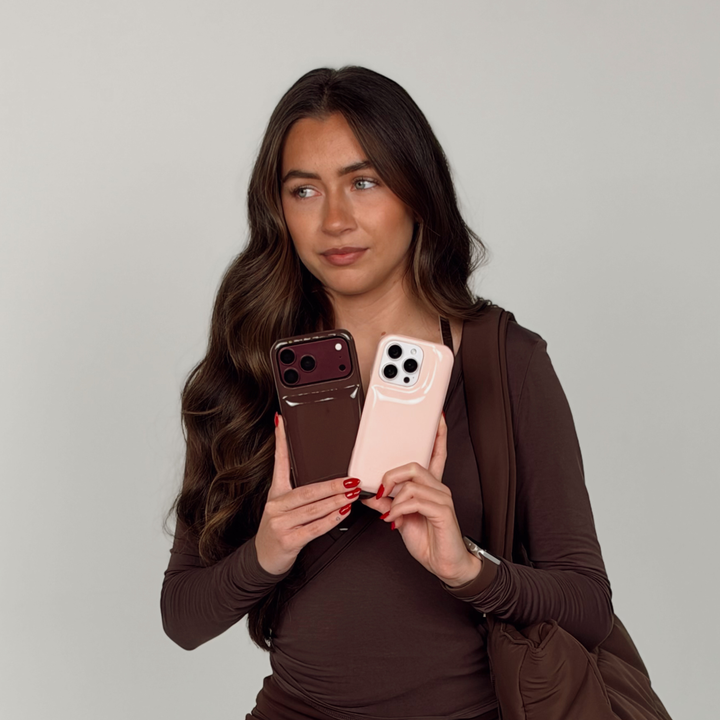 Woman wearing a brown pilates outfit holding two iphones. One is in a glossy chocolate brown Coconut Lane puffer phone case with embossed logo. The other iphone is in a pastel pink Coconut Lane puffer phone case.On her wrist is a Coconut Lane NAKD Chocolate brown apple watch strap. 