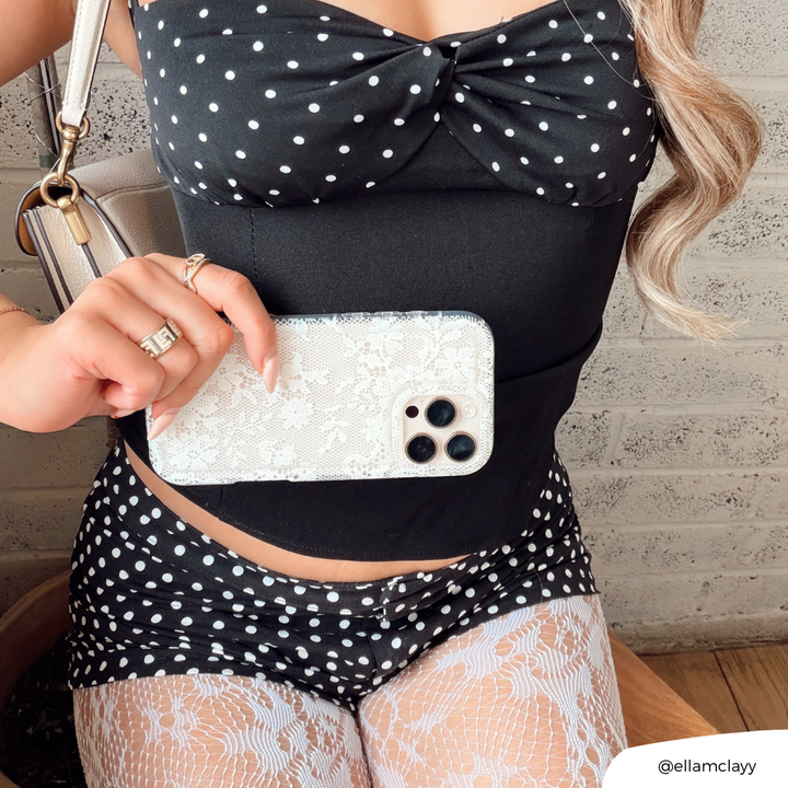 Woman in polka dot outfit and lace tights holding Coconut Lane clear phone case with white lace design by a brick wall.