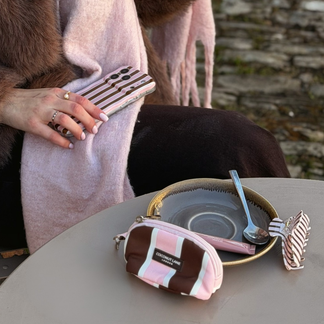 Coconut Lane Cocoa & Pink Stripe mini keyring bag featuring chocolate brown, baby pink and white stripes, placed on a table alongside a coffee. In the image is the matching claw clip and wavy phone case.
