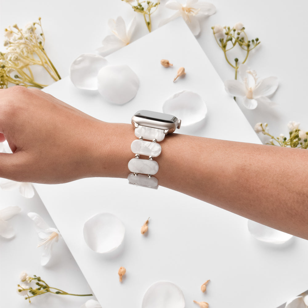 A person wearing the stylish White Pearl Beaded Apple Watch Strap by Coconut Lane, featuring a beautiful white patterned design with pearl-like beads. The wrist is angled so that the watch face is turned away. The background is adorned with scattered white flower petals and small white blossoms on a pristine white surface.