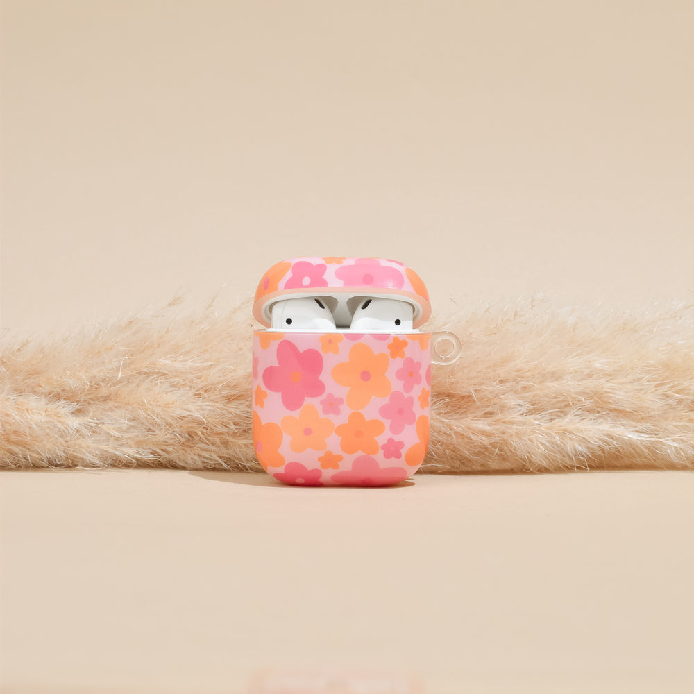 The Coconut Lane AirPods Case - Pink & Orange Bloom is displayed against a beige background, housing a pair of white wireless earbuds. Utilizing IMD Print Technology, the vibrant and durable floral design enhances the case's appeal. The lid is open, revealing the earbuds inside, while pampas grass arranged behind adds a decorative touch.