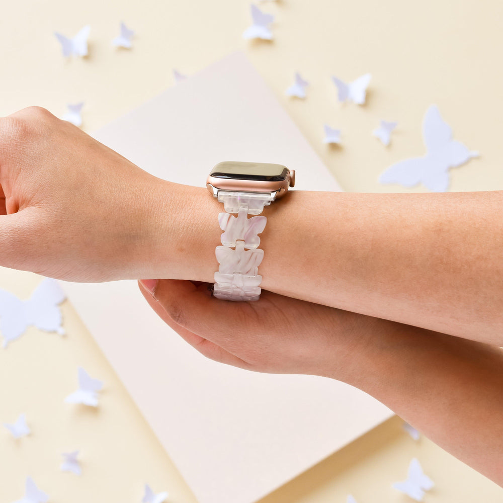 A person’s wrist adorned with the Luxe Holo Butterfly Apple Watch Strap by Coconut Lane, showcasing its white band with elegant butterfly patterns. The background highlights holographic butterflies scattered across a light surface. In a close-up pose, the person’s hand prominently displays the adjustable strap.