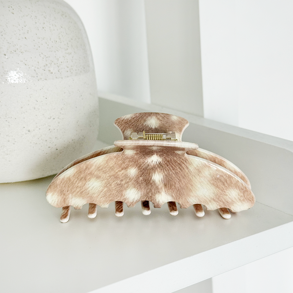 Hair claw clip with a brown and cream mottled animal-print pattern, resting on a white shelf next to a textured ceramic vase.