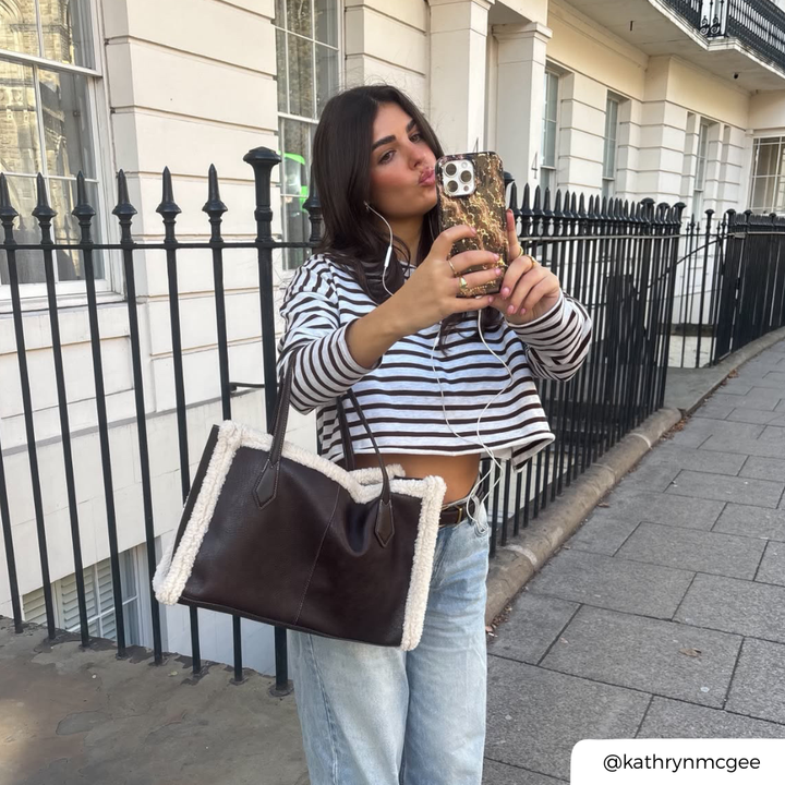 A woman with blue jeans and a striped long sleeve top, holds an iphone in a beige and dark brown marble design with subtle gold glitter flecks phone case.