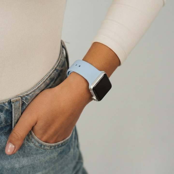 A smartwatch with a square face and a light blue silicone strap, specifically the NAKD Apple Watch Strap - Sky Blue by Coconut Lane, is displayed against a white background. The black watch face, adorned with white hands, and the Sky Blue strap, featuring a silver buckle and multiple adjustment holes, evoke spring-inspired pastels.