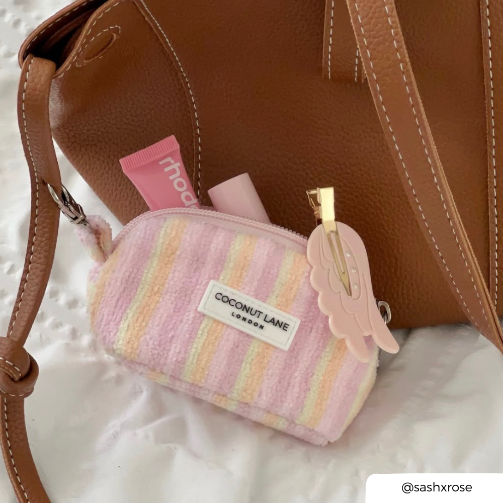A pastel striped zippered pouch with a white label reading 'COCONUT LANE' and a pink angel-shaped clip, styled next to a brown handbag on a white textured surface.