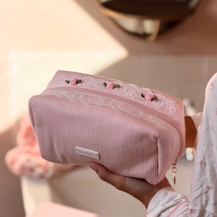 A person holds a light pink cosmetic pouch with a textured fabric body, a white lace trim, and embroidered pink roses on top.
