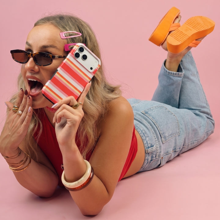 A woman lying on her stomach against a solid pink background, holding an iPhone with a red and orange striped case. She is wearing sunglasses, a red top, light-wash blue jeans, and bright orange platform shoes.