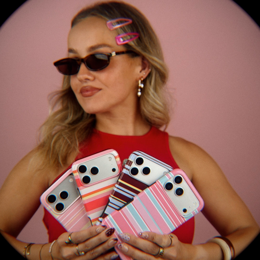 A fish eye effect image of a woman wearing retro sunglasses and old school hair clips holds four striped iPhone cases against a pink background