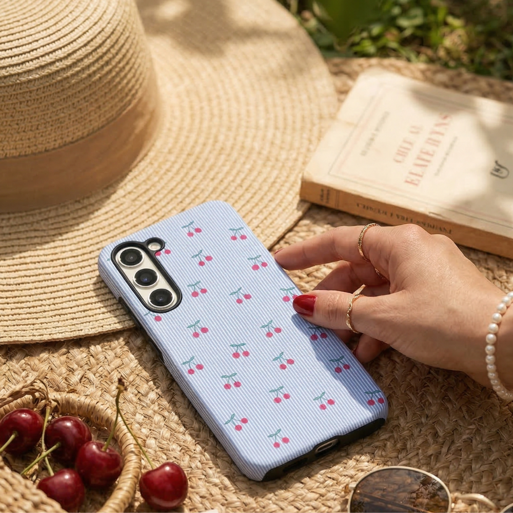 Phone case with cherry pattern held by a hand, surrounded by a straw hat, book, and cherries on a woven surface.