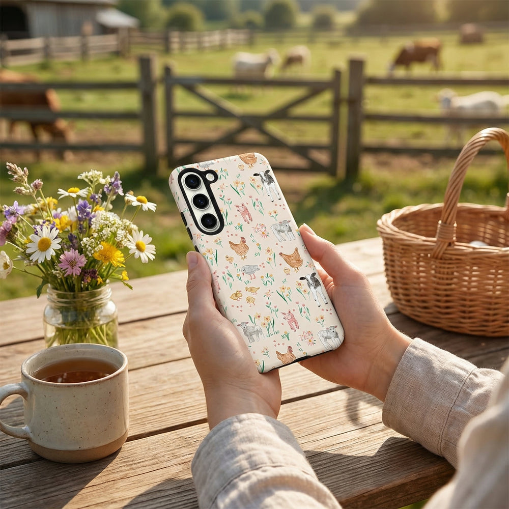 Person holding a phone with a farm-themed case, sitting at a table with a cup of tea, flowers, and a basket in a pastoral setting.