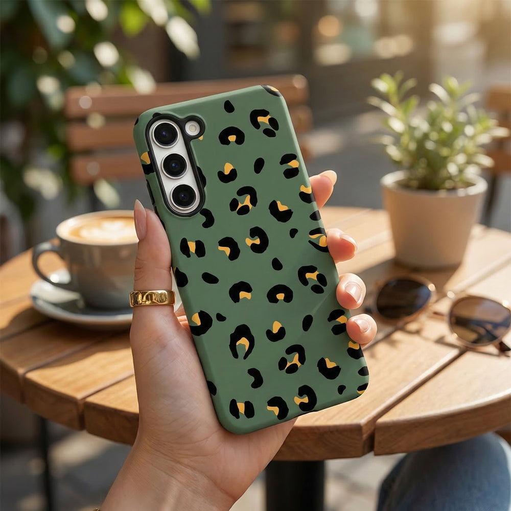 Hand holding a khaki leopard print Samsung Tough Phone Case at an outdoor café table with coffee and sunglasses.