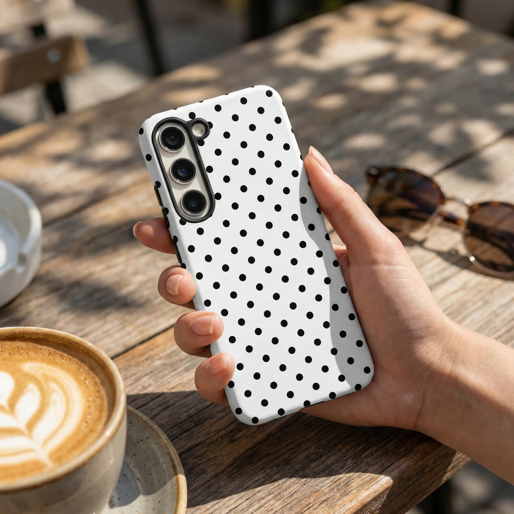 Hand holding white polka dot Samsung phone case by Coconut Lane beside sunglasses and a latte on a table.