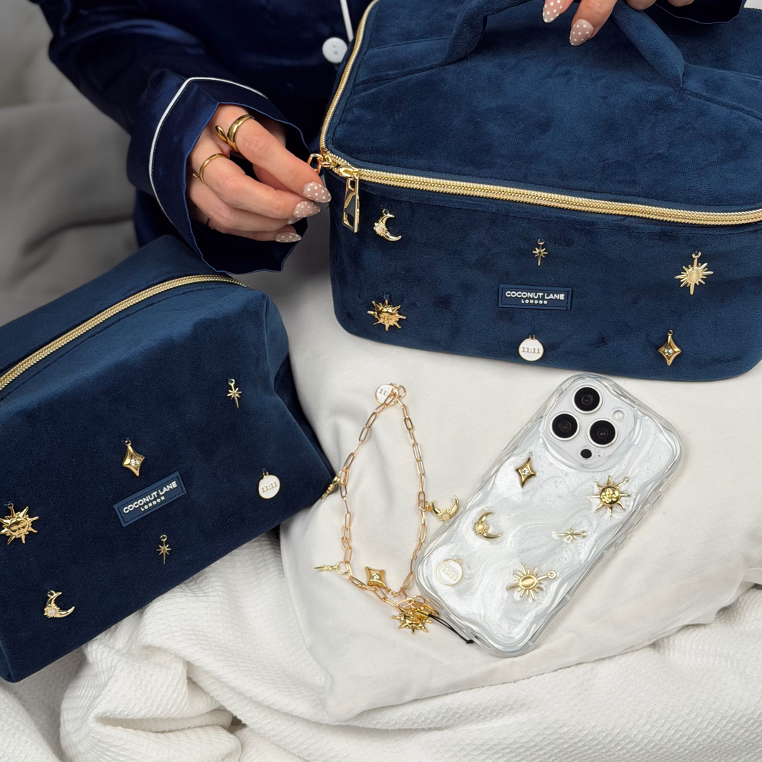 Woman wearing satin navy pyjamas with  a phone in a stardust after dark phone case with gold charm embellishments. The phone has a matching gold chain phone strap with celestial style charms, including a moon, sun, star, diamond shape and 11:11 charm. 

In the photo is the matching makeup bag and vanity case with navy velvet material and gold charm embellishments.