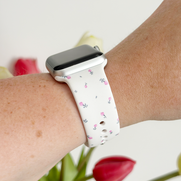 A person's wrist sports the Ditsy Floral White Apple Watch Strap by Coconut Lane, featuring a pink floral pattern against a white band. The background showcases blurred tulips in vibrant red, white, and pink shades, enhancing the scene's colorful ambiance.