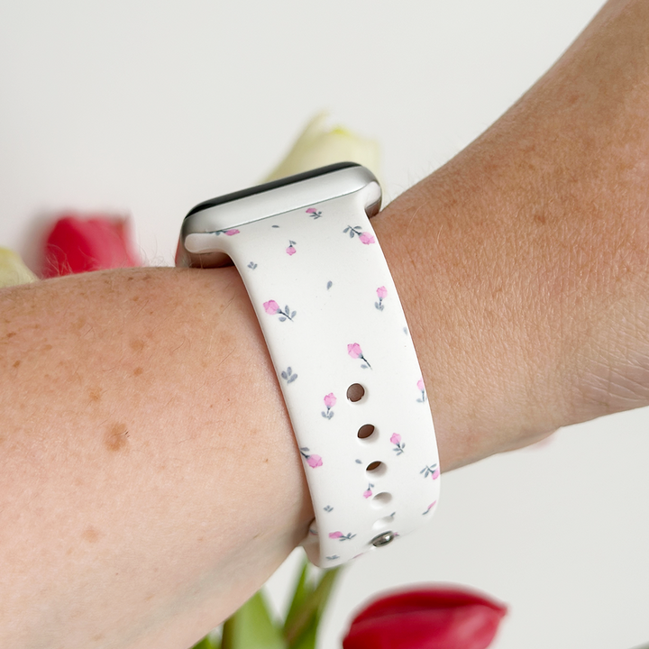 A close-up of a person's wrist wearing a white smartwatch with the Coconut Lane Ditsy Floral White Apple Watch Strap, adorned with small pink flowers. The background is blurred out but shows some red and white flowers.