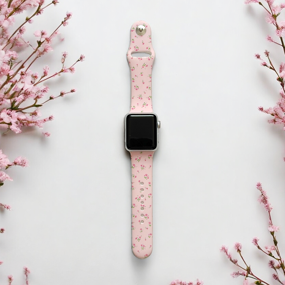 Pink floral Apple Watch strap with a ditsy pattern displayed on a white surface, pink flowers alongside.