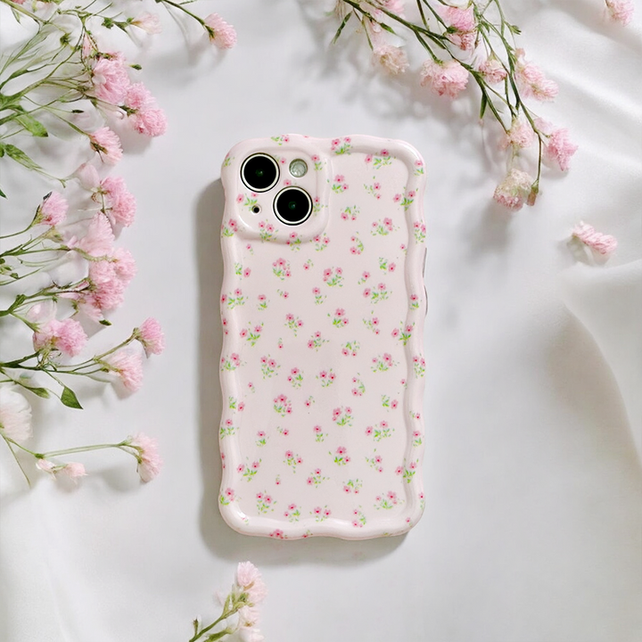 A smartphone with the Wavy Phone Case – Ditsy Floral Pink by Coconut Lane lies on white fabric. Small pink flowers encircle the phone, enhancing the scene with a soft, elegant touch.