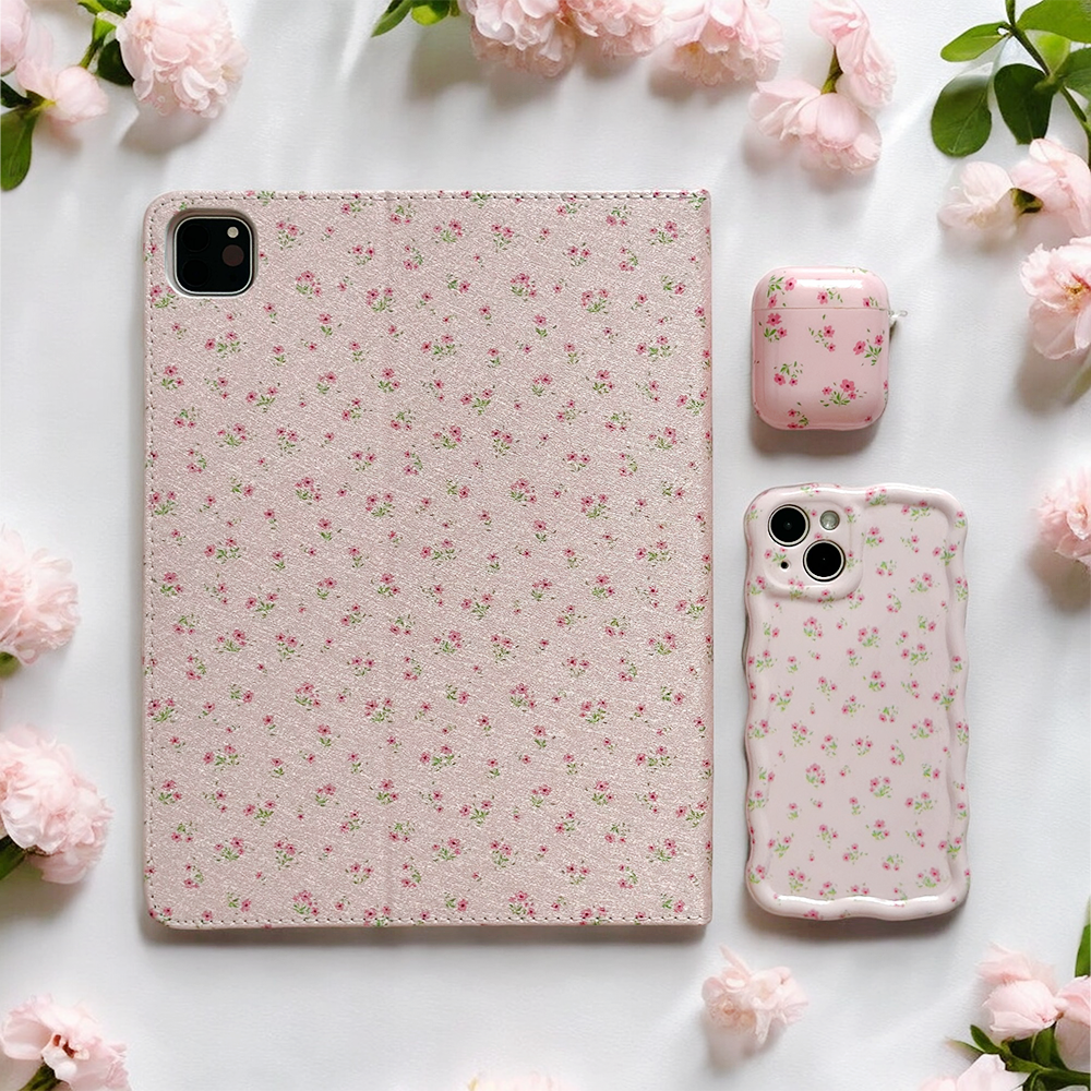 A Coconut Lane Ditsy Floral Pink iPad case, complemented by matching iPhone and AirPods cases, lies on a white surface surrounded by pink flowers. The delicate print adorns each vegan leather item, creating a cohesive and aesthetically pleasing arrangement.