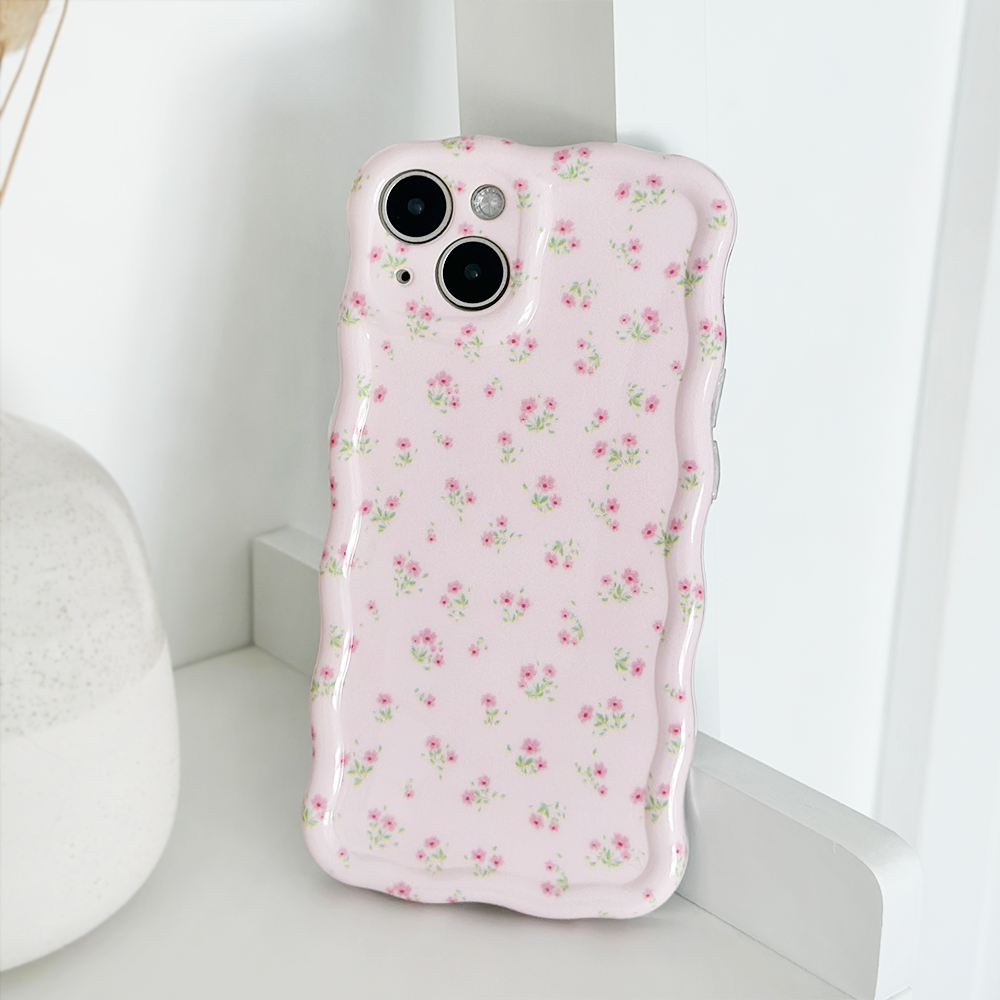 A close-up of a smartphone encased in the Wavy Phone Case - Ditsy Floral Pink by Coconut Lane, featuring delicate ditsy floral patterns in pink. The phone is propped up against a white wall next to a white vase, creating a clean and minimalist aesthetic.