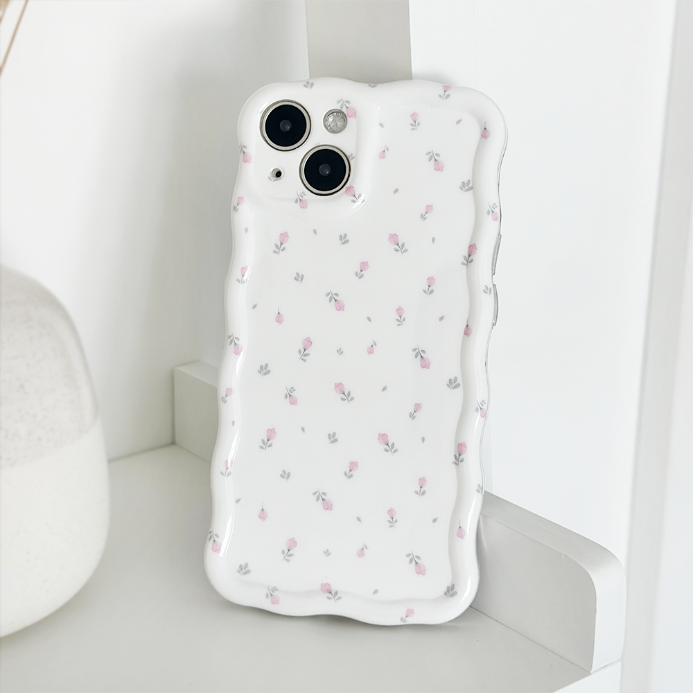 The image features a white smartphone encased in a Wavy Phone Case - Ditsy Floral White by Coconut Lane, offering extra protection and a decorative touch with its subtle floral pattern. The phone, equipped with dual cameras, is leaning against a white textured object on a smooth white surface.