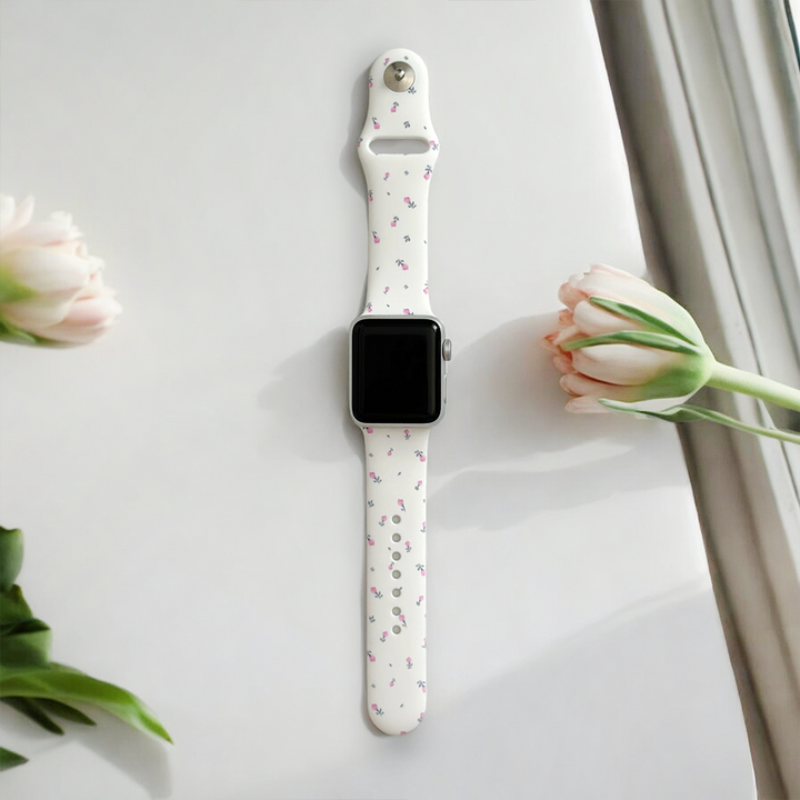 A smartwatch with a Ditsy Floral White Apple Watch Strap by Coconut Lane, featuring pink and green patterns, is placed on a light-colored surface near a window. Delicate pink tulips on either side of the watch add a touch of elegance to the scene.