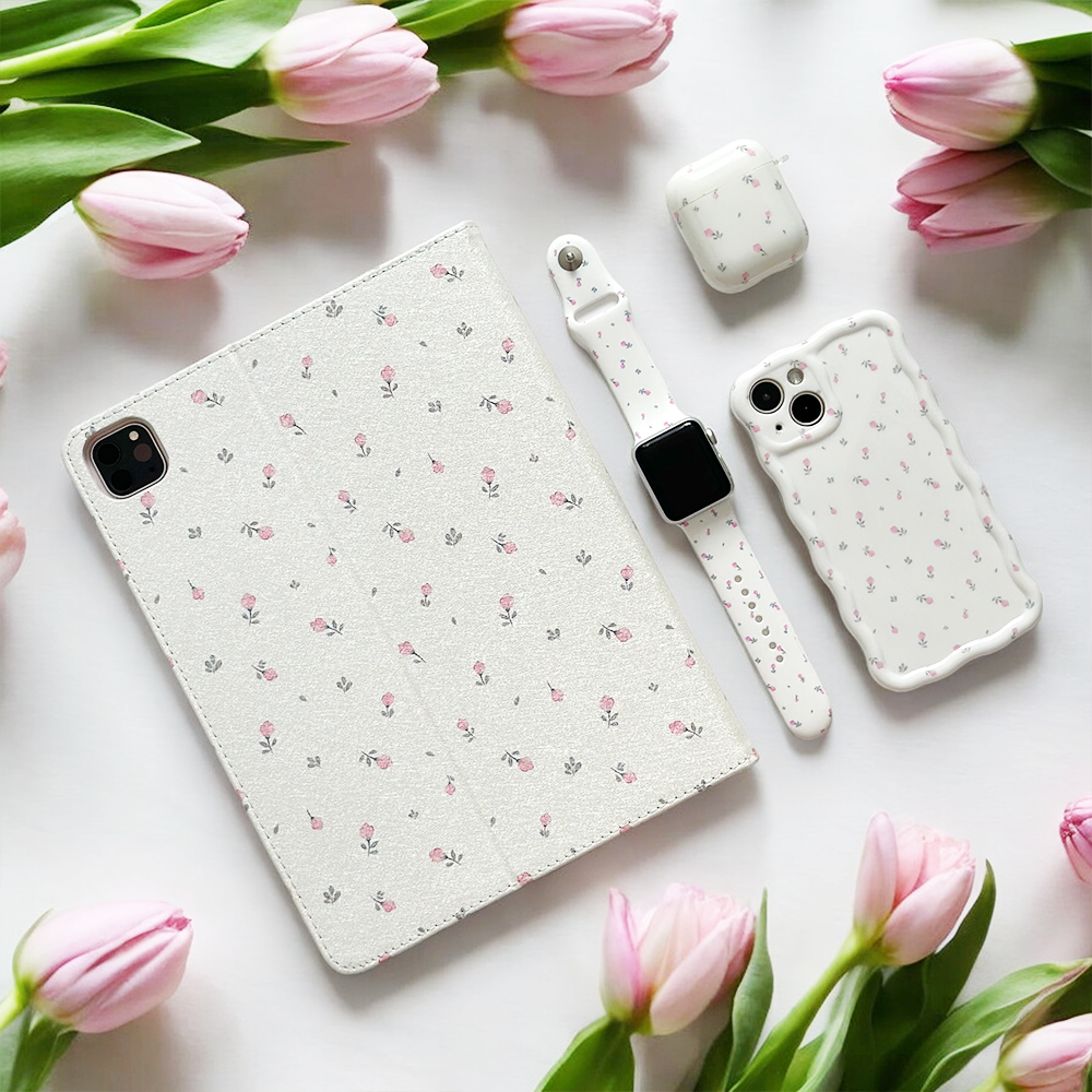 A neatly arranged flat lay features a Coconut Lane Ditsy Floral White iPad Case, smartwatch, smartphone case, and wireless earbuds case, all adorned with a delicate floral pattern. The vegan leather accessories are beautifully surrounded by fresh pink tulips on a white background.