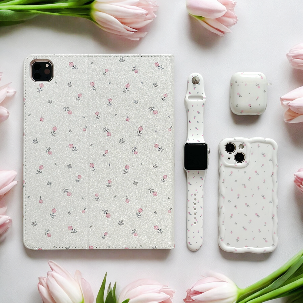 A flat lay image featuring a tablet case, smartphone case, wireless earbud case, and the Ditsy Floral White Apple Watch Strap by Coconut Lane. All accessories have a matching white and pink floral pattern. The arrangement is surrounded by light pink tulips, creating an aesthetically pleasing composition.