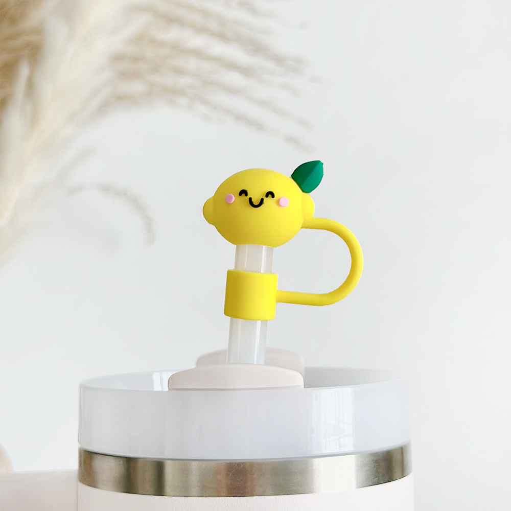 A close-up of a drink cup featuring a clear lid and a metal band around its base. The adorable Happy Lemon Straw Cover by Coconut Lane, showcasing an endearing lemon character with pink cheeks and a green leaf, serves as an attractive high-quality silicone straw topper above the cup. The background is white with soft-focus details.