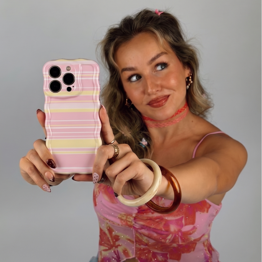 A woman taking a selfie with a phone case featuring a pastel pink, yellow, and white wavy stripe pattern. She is wearing a pink patterned top and various bracelets.