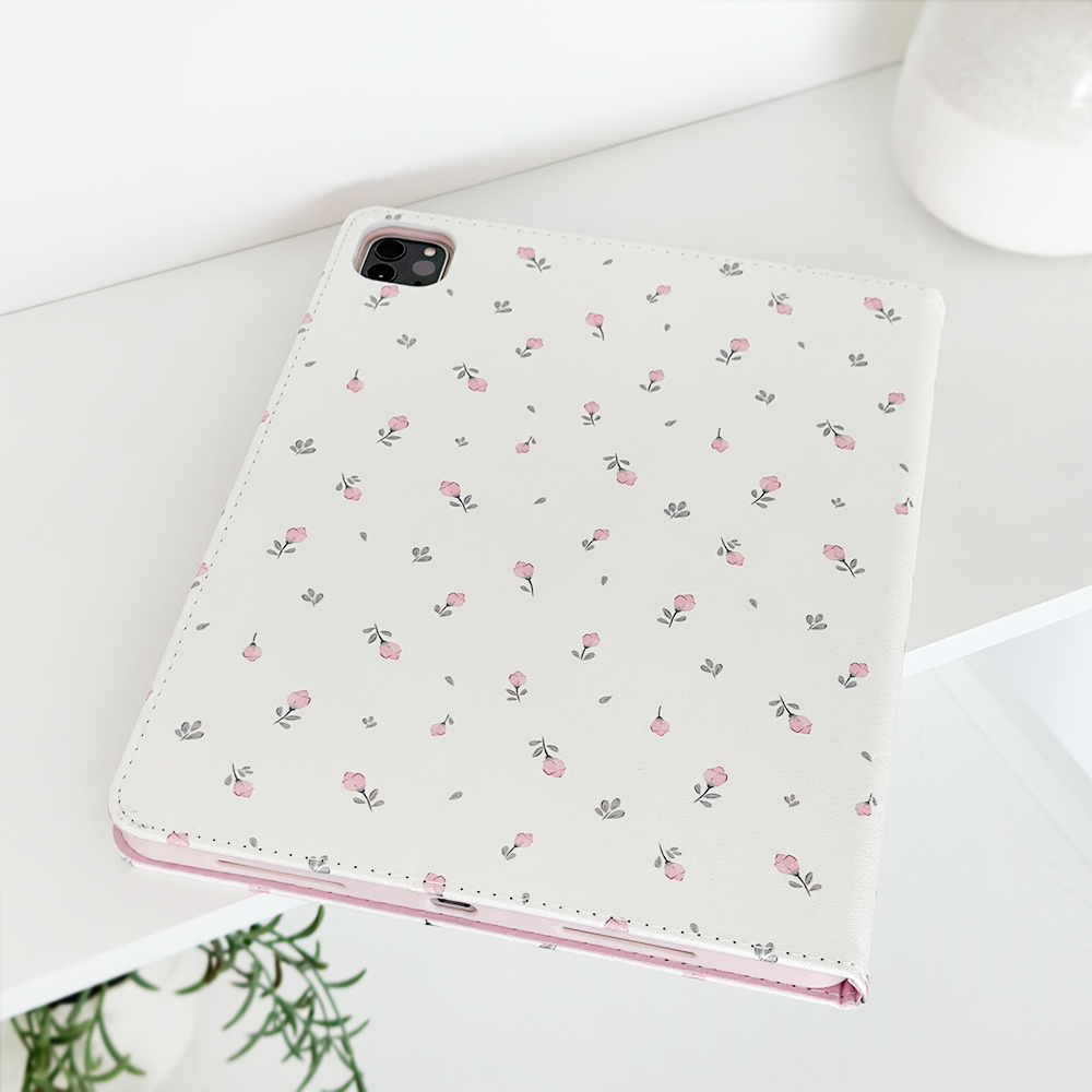 A Coconut Lane Ditsy Floral White iPad Case, adorned with a delicate pink flower pattern, is placed on a white surface. The tablet camera is visible through a cutout in the vegan leather case. A small green plant and a white vase in the background add a touch of greenery to the minimalistic scene.