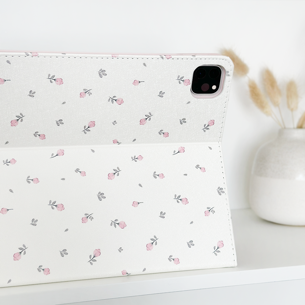 A Coconut Lane Ditsy Floral White iPad Case, adorned with delicate pink roses and subtle gray leaves, stands upright on a white surface. To its right, a white vase holds dried beige flowers. The vegan leather cover of the case not only provides anti-slip properties but also seamlessly merges style and functionality against the simple white backdrop.